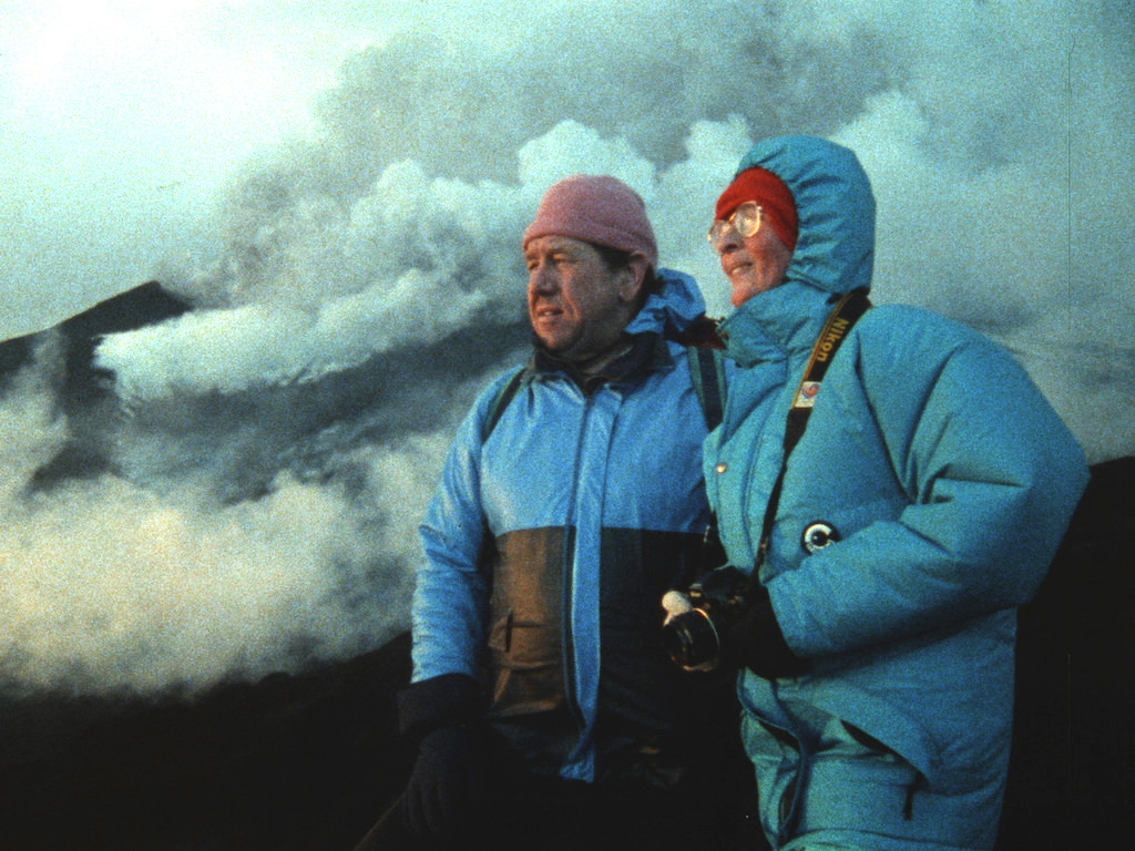 Katia and Maurice Krafft, in blue winter jackets, gaze upon a volcano in the distance as smoke, steam and ash swirl behind them. (Credit: Image'Est)